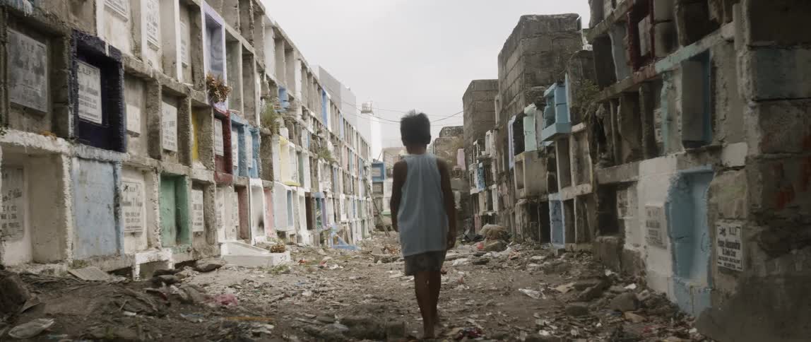 A little boy walks barefoot on dirt and debris through two walls of cement cubes