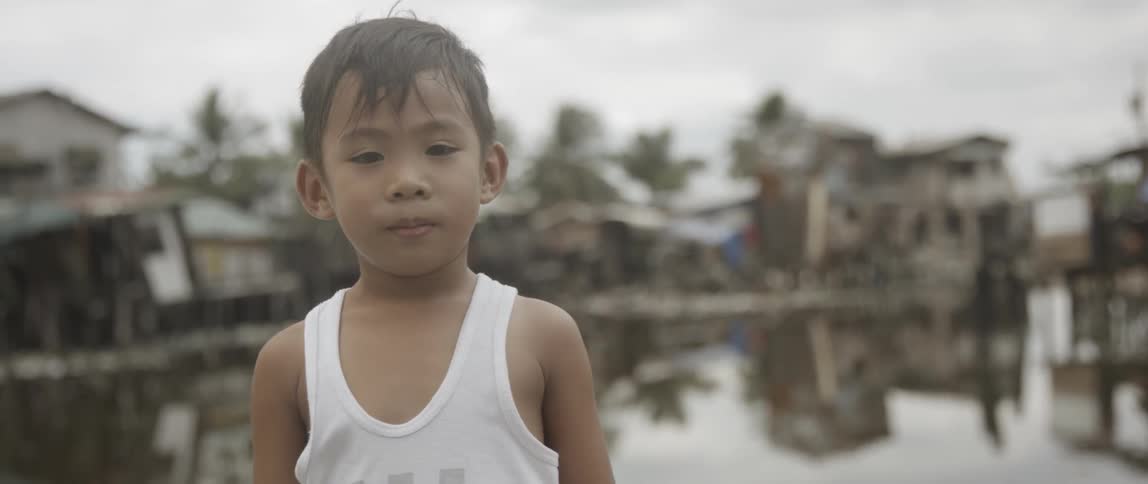 A boy in a tank top standing in front of a village on stilts while looking at the camera