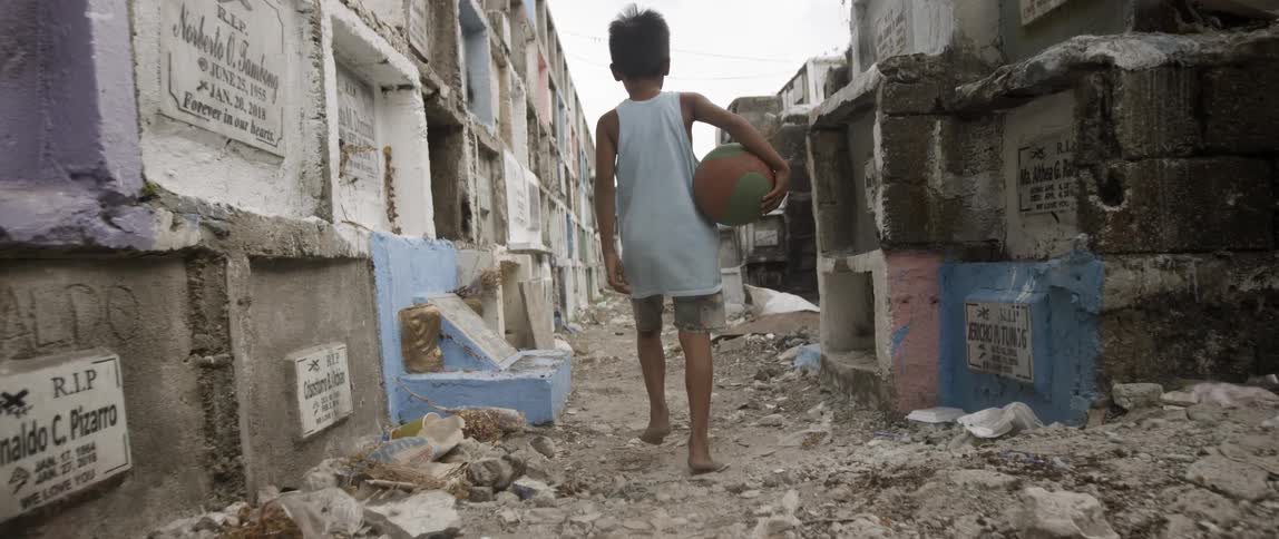 A young child carries a basketball through a dirt town road