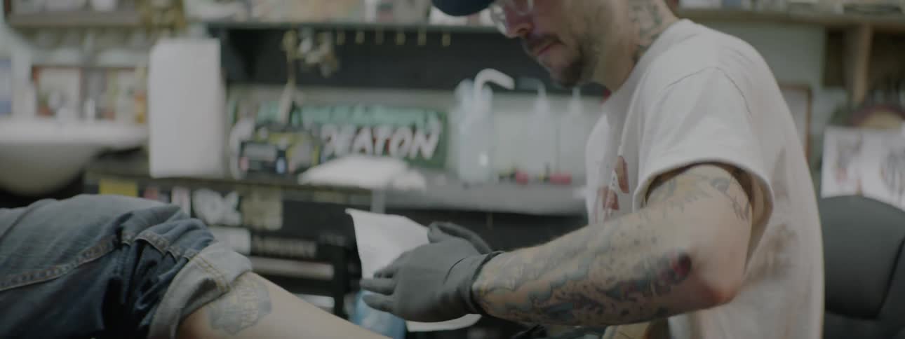 Tattooed man wearing cap and glasses rubs another mans tattooed leg indoors