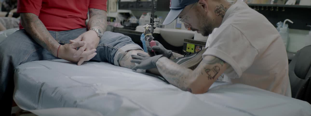 A male tattooist is tattooing the lower left leg of a white male