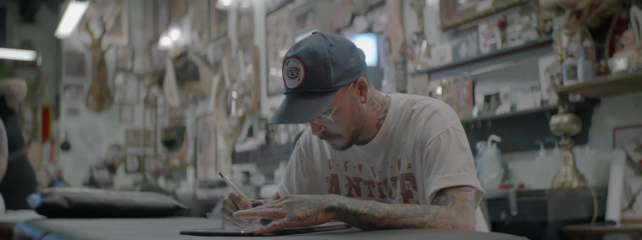Heavily tattooed man works on drawing in tattoo shop