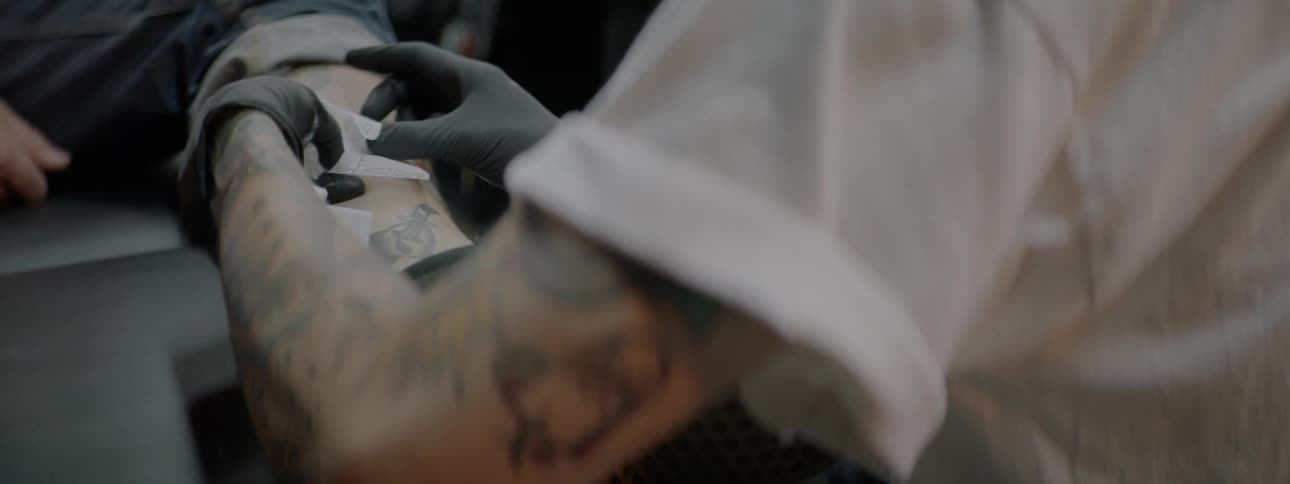 A white man with a tattoo prepares to tattoo another person's leg