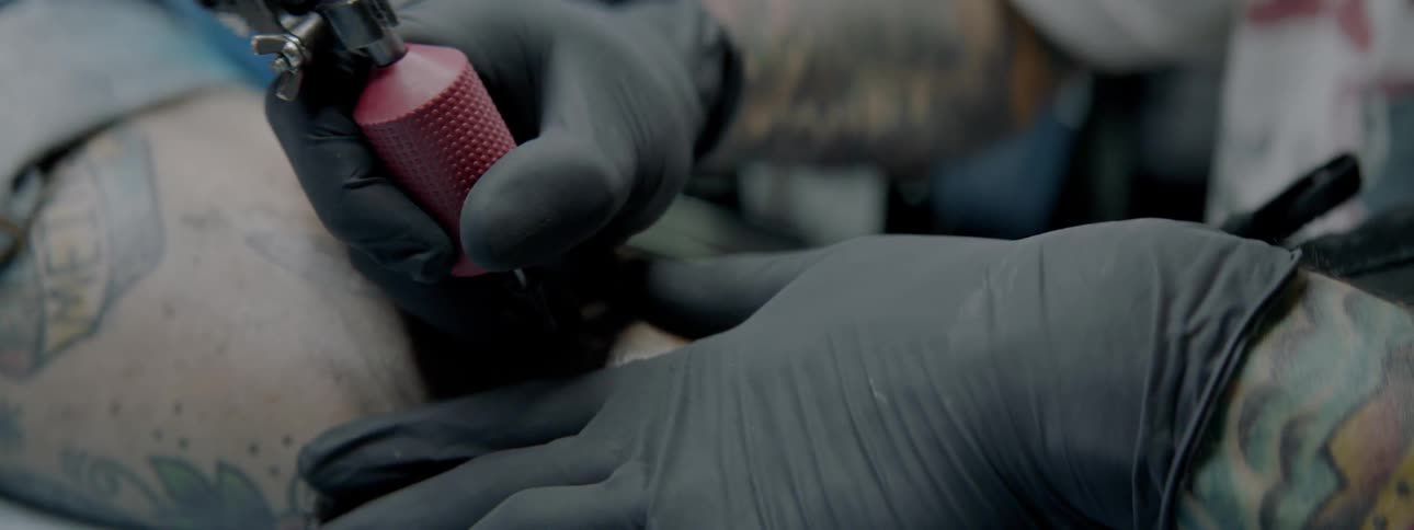 A tattooist tattoos on a man while wearing black gloves
