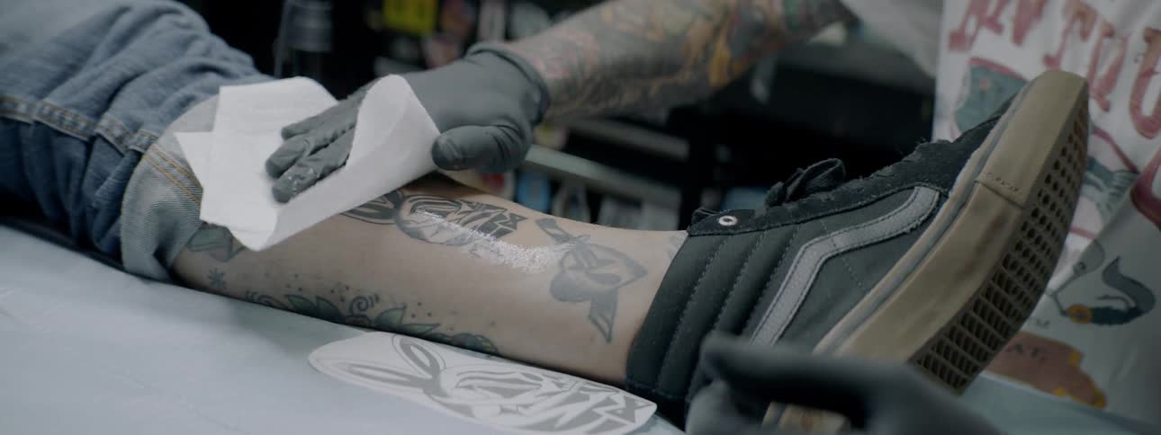 Man cleans another mans tattooed leg on the stretcher indoors