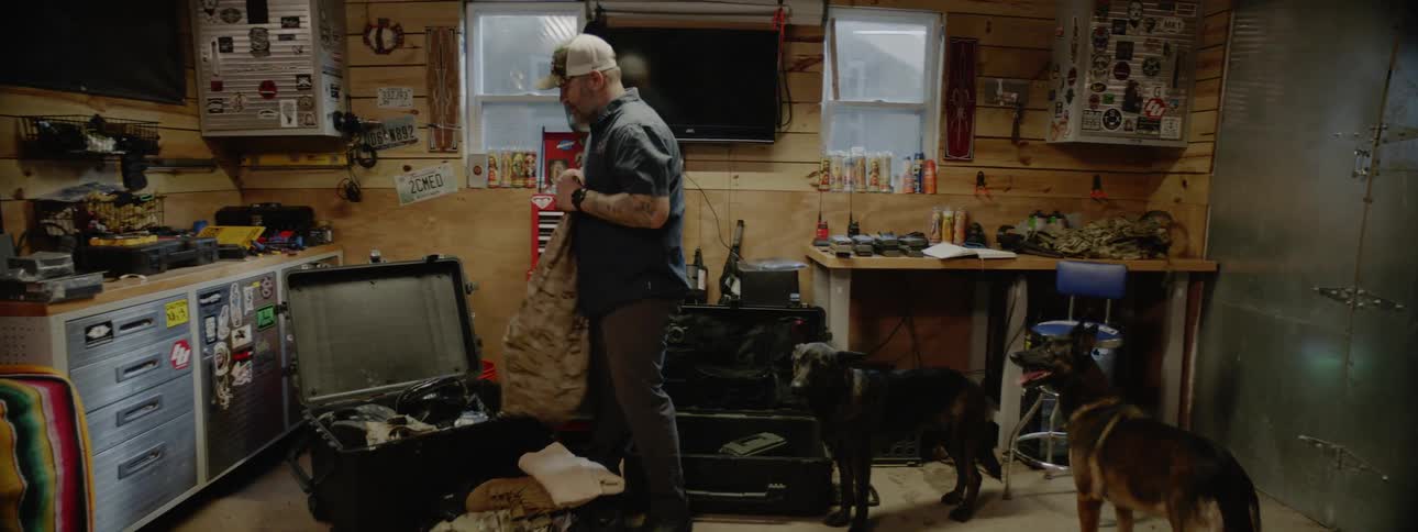 A man is in his garage with two dogs and he is getting pants out of a trunk