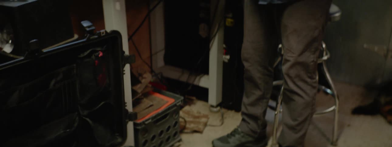 A white man stands over a case of military gear while taking a knife in and out of a sheath