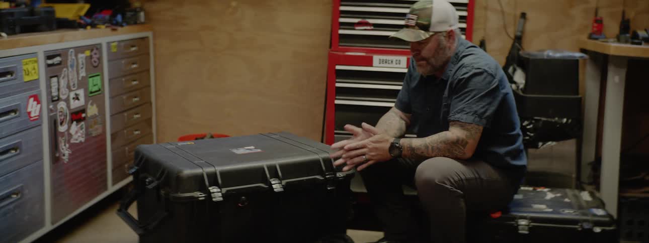 An old man in a ball cap puts military clothing in a plastic case and then pets a brown dog
