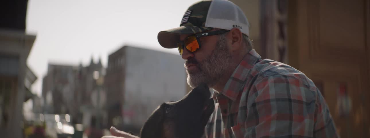 A man kneeling on the sidewalk petting his dog who is licking his face