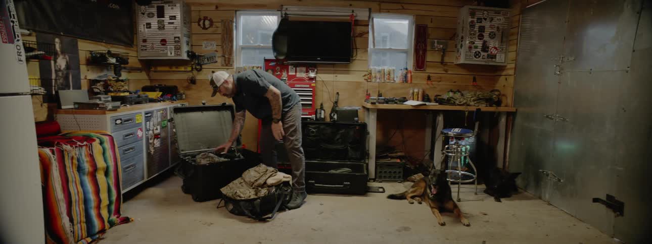 A man sitting at a desk reading a book before rising and packing clothing in a trunk