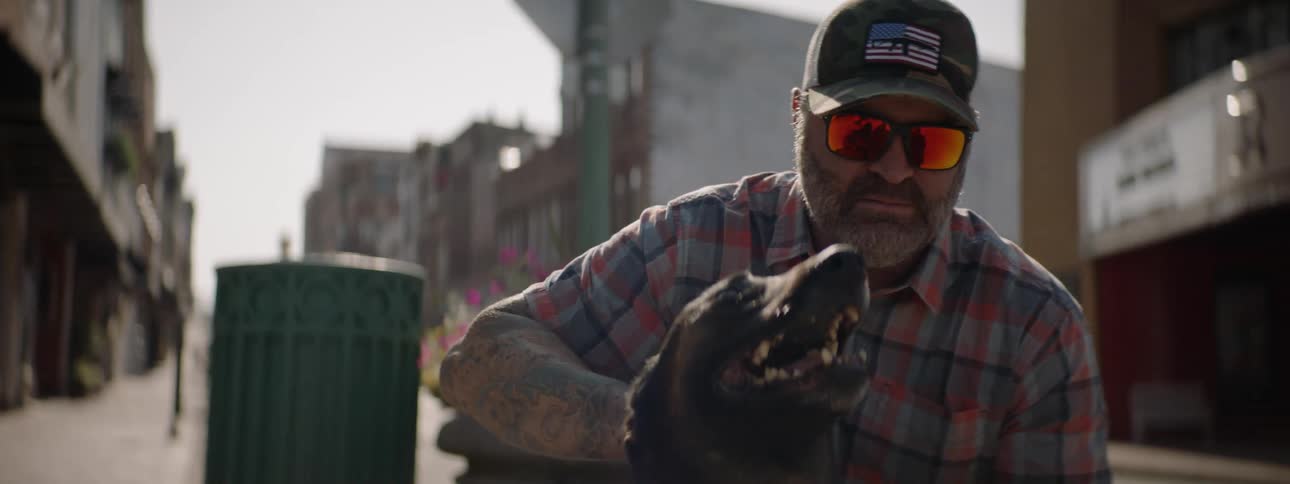 A man wearing sunglasses with reflective lenses pets his dog on a sunny sidewalk