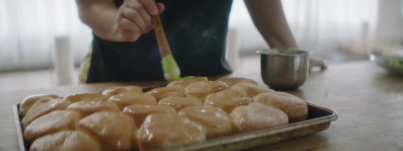 A person is using a brush to apply a glaze to the tops of uncooked rolls already on a baking pan