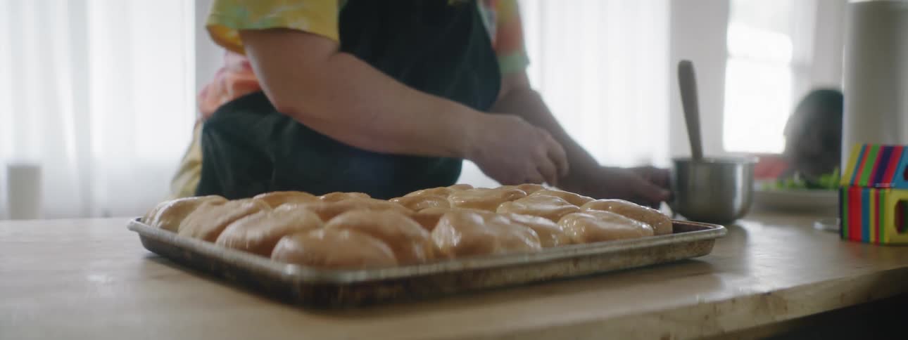 Woman wearing black apron brushes egg over pastries on the table while black boy looks at it at home in the daytime