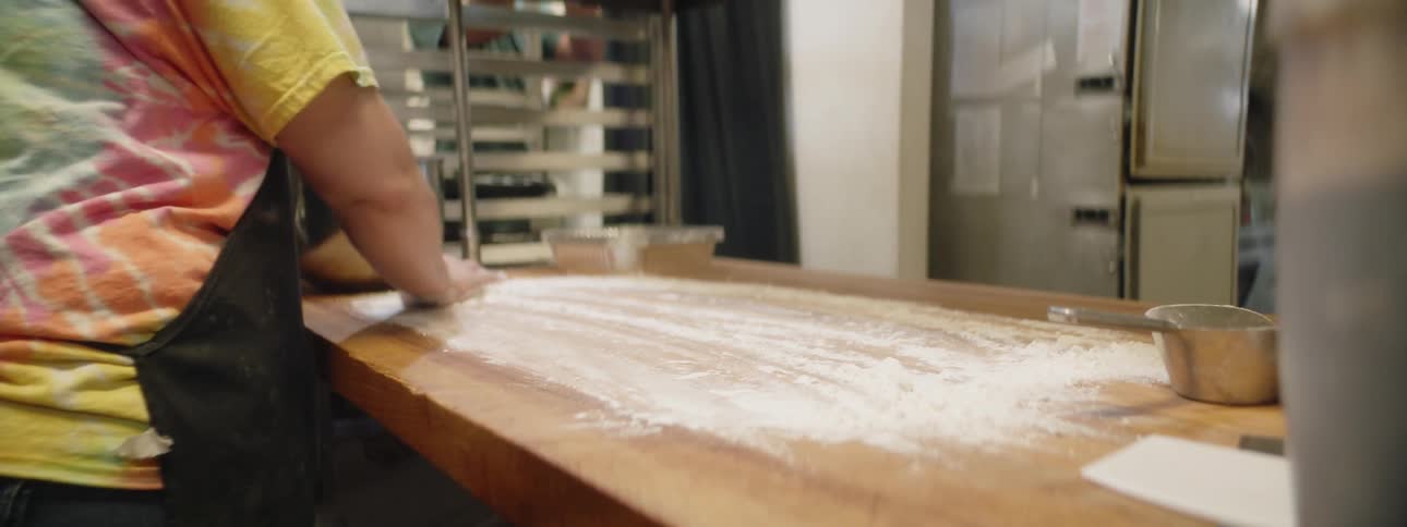 A woman in an apron and tie dyed shirt spreads flour on a wood counter in a commercial kitchen