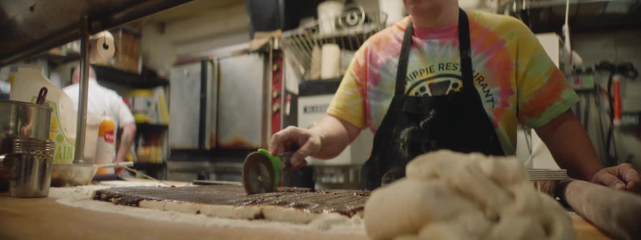 Woman in tie dye shirt cuts cinnamon bread with pizza cutter in commercial kitchen