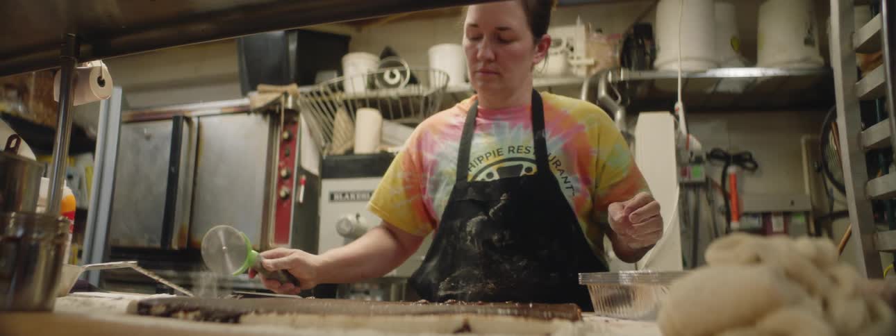 A woman in an apron cuts cinnamon rolls in strips in a commercial kitchen