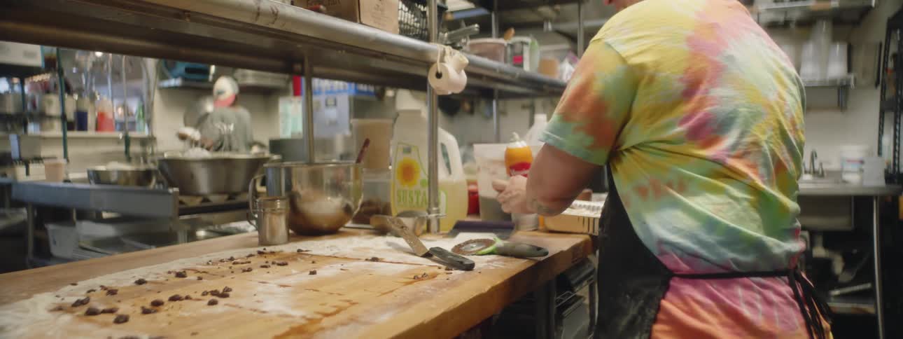 A baker places a cinnamon roll in a pan then starts to clean off his butcher block counter