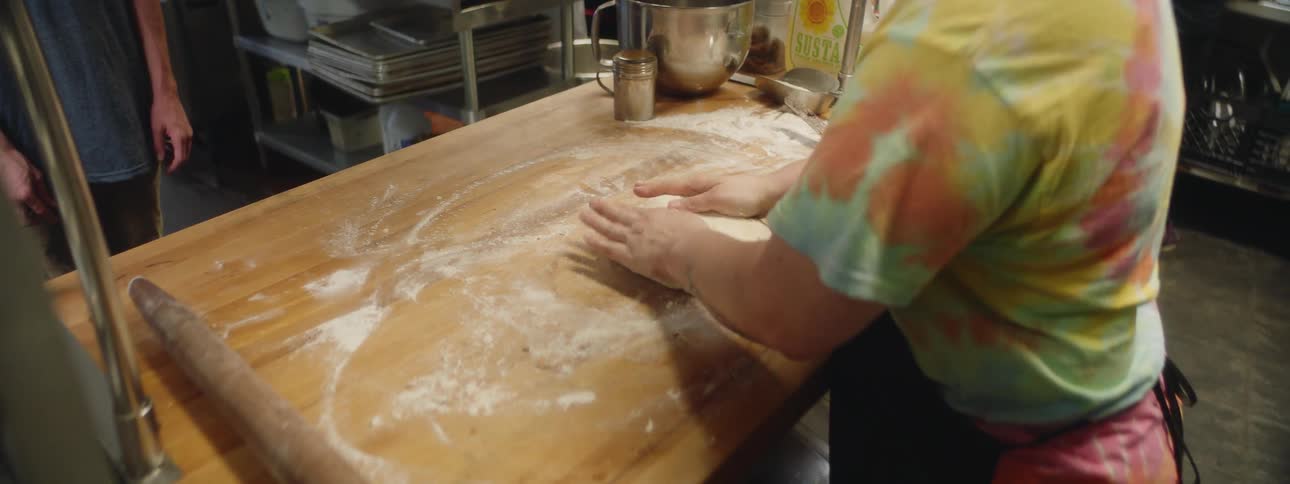 Woman wearing black apron rolls dough with rolling pin on the wooden board indoors