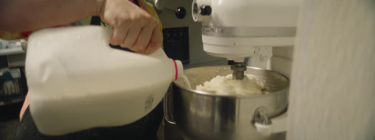 A man in an apron adds milk to a commercial mixing bowl filled with mashed potatoes in a restaurant kitchen