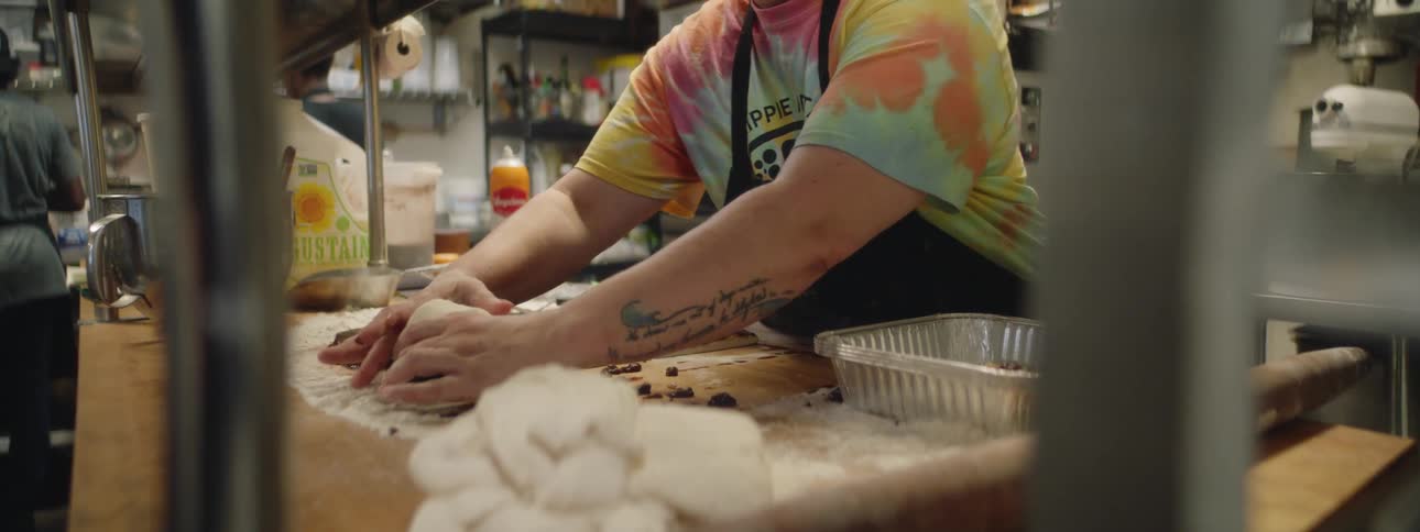 A man in an apron and tie dyed shirt prepares cinnamon rolls in a commercial kitchen
