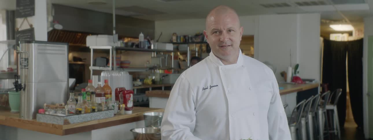 Cook holds a bowl of salad in hand and smiles in the kitchen during the day