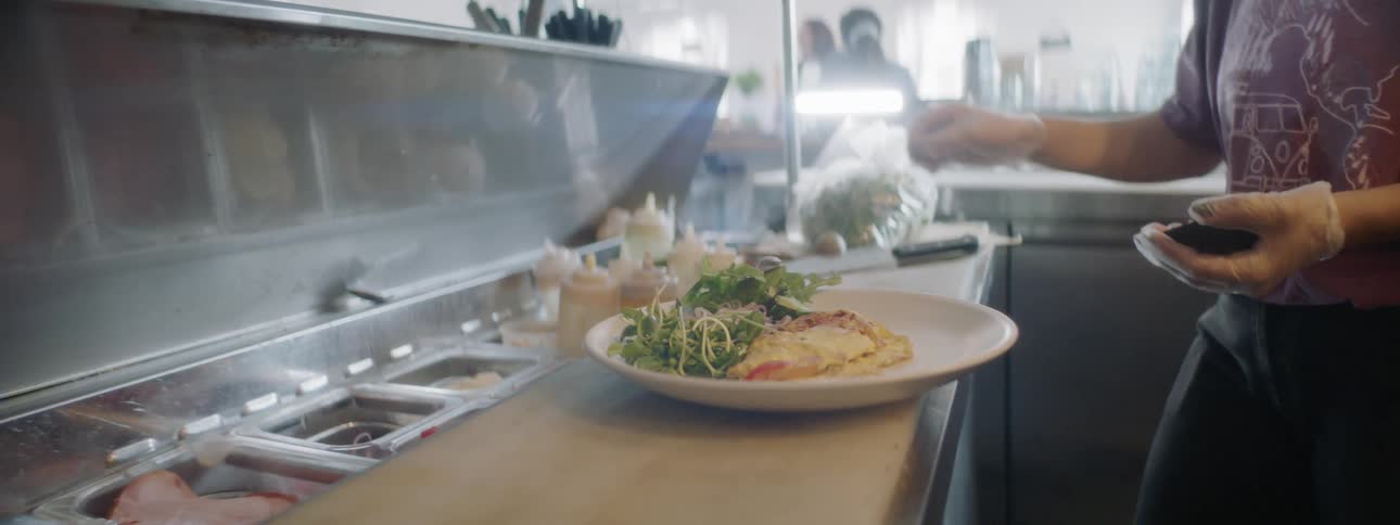 A person in a restaurant kitchen is holding half of an avocado in their hand swings a large knife downward at the pit to chop into it and remove it as they prepare and plate an egg omelet