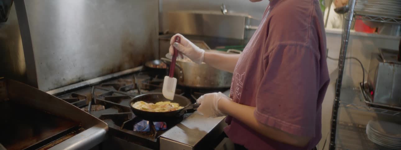 Woman stirs food in skillet with spatula over gas stove in commercial kitchen