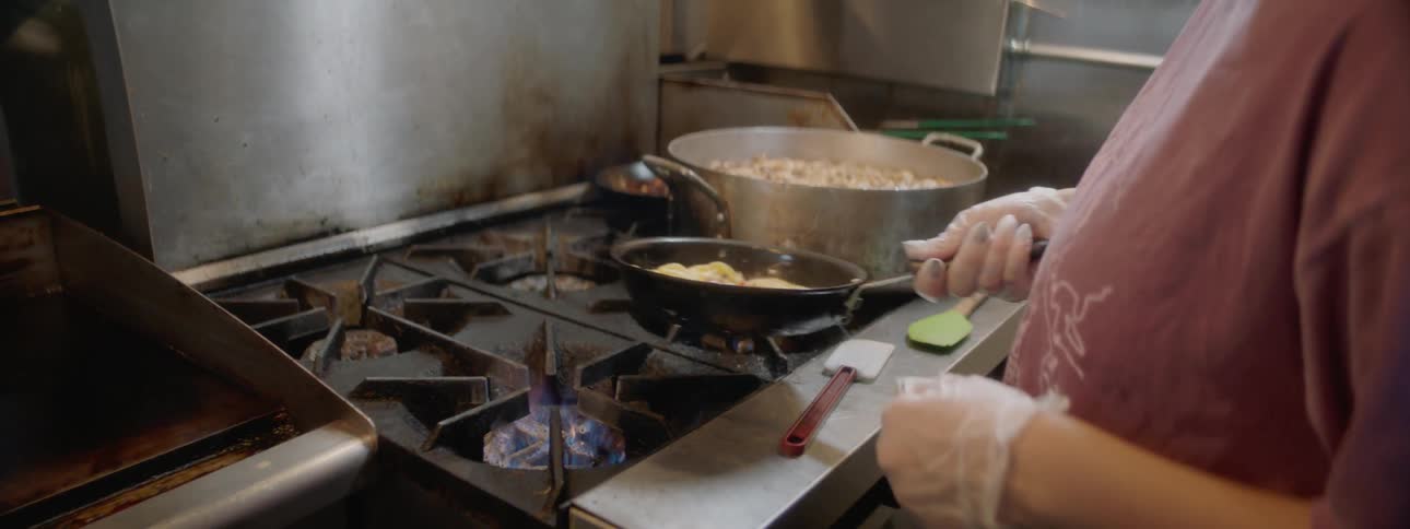 A woman is in a restaurant kitchen cooking with a skillet and flipping the food in it