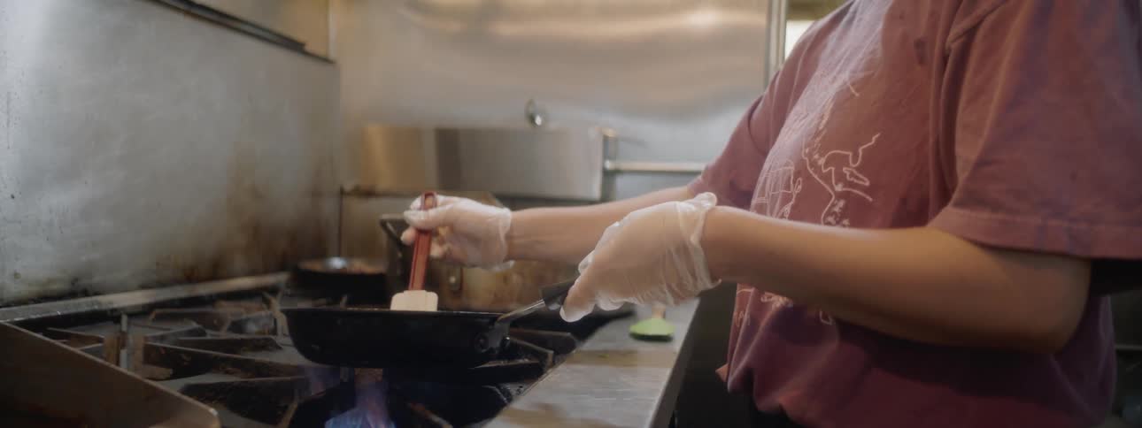 A white female is cooking over a large commercial stove in a restaurant kitchen