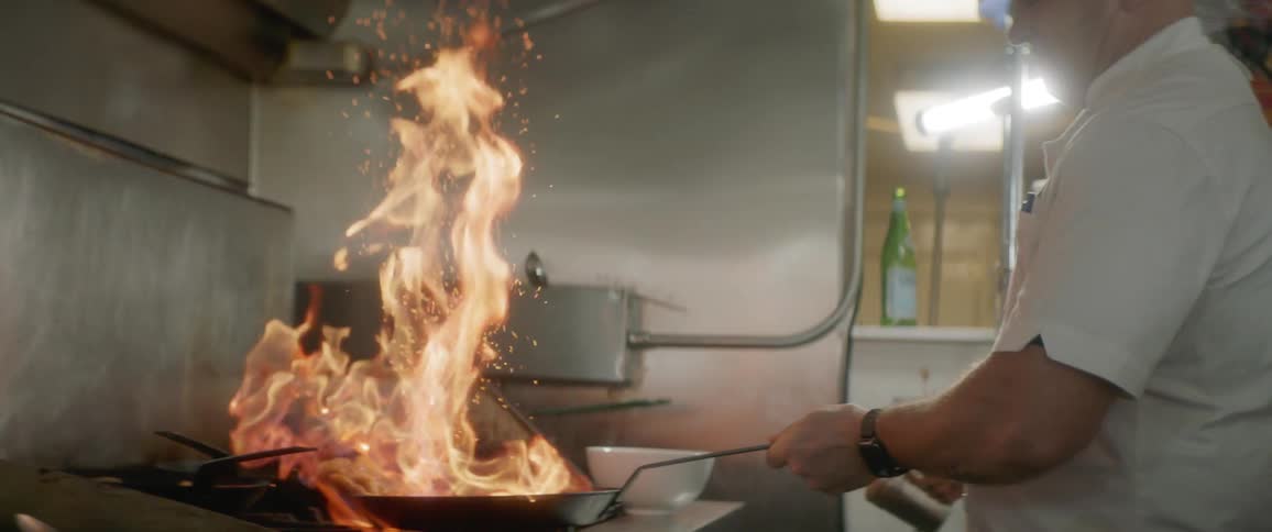 A chef in a commercial kitchen adds seasoning to a pan that has a flame rising from it