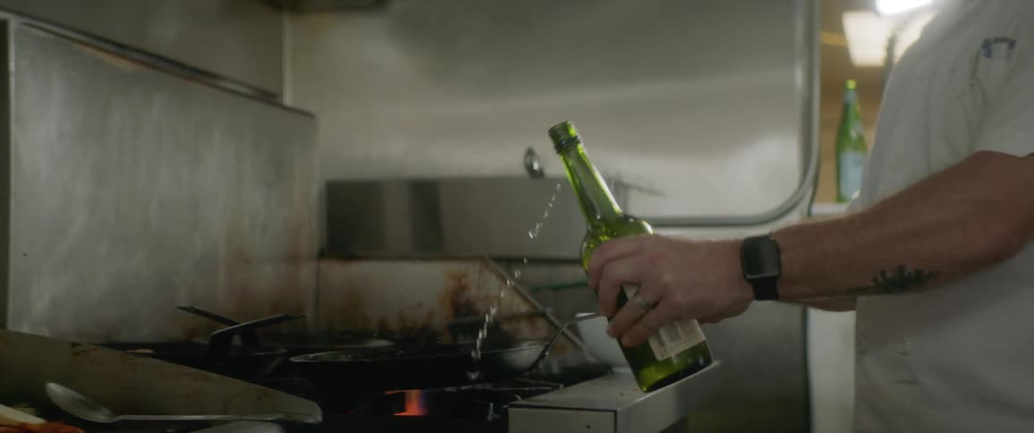 A cook pouring wine into a pan causing flame to flare up from the burner
