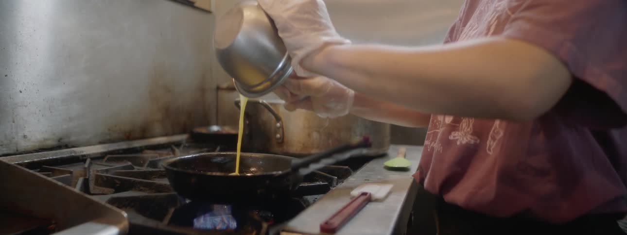 A young woman stirs eggs and adds them to a pan
