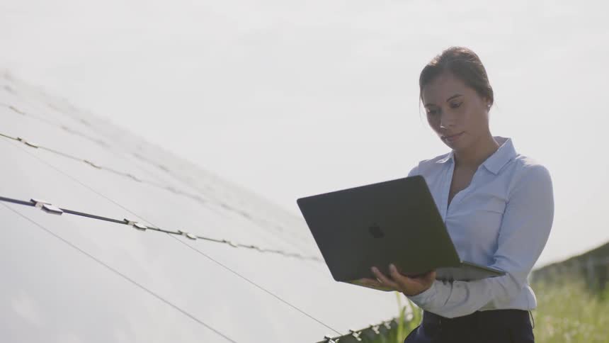 A woman standing next to a row of solar panels opens up a laptop