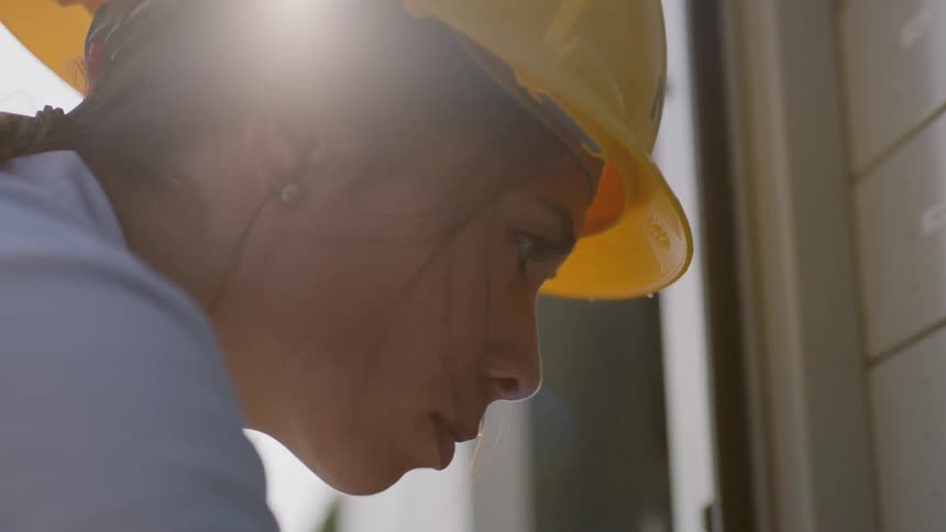 A woman in a yellow hard hat and carrying a clipboard is focused very intently as she is inspecting a piece of equipment