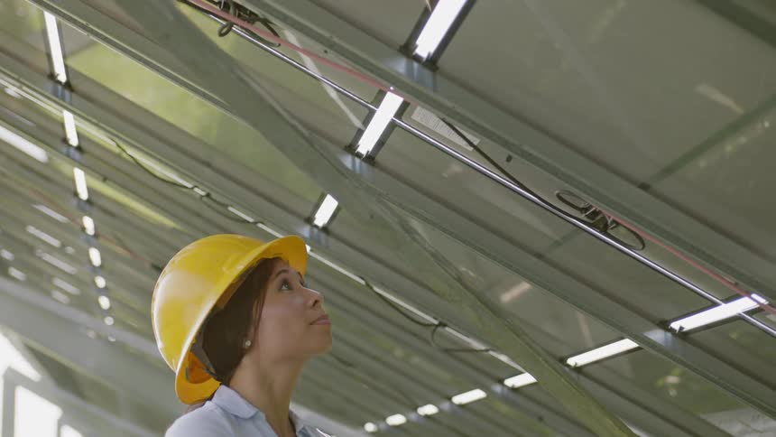A woman is standing under a row of solar panels to record a model number while typing on a laptop