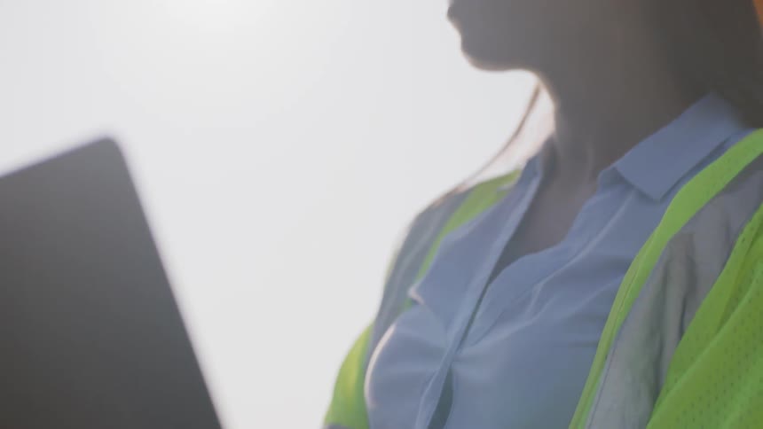 A woman wearing a helmet and high visibility vest types on a laptop