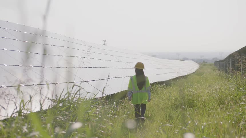 A woman in a hard hat and safety vest walks next to a row of solar panels