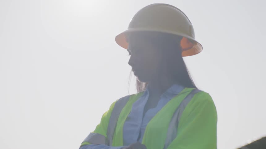 A woman wearing construction shirt and helmet works on a laptop in the field