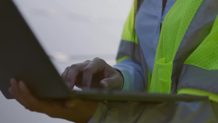 A woman wearing a hardhat and safety vest holds and taps on a laptop