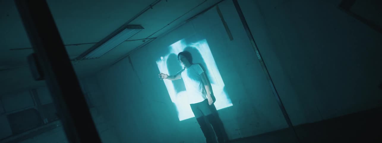 A man stands against a wall with an arm outstretched while lights and images are flashed over him and the wall