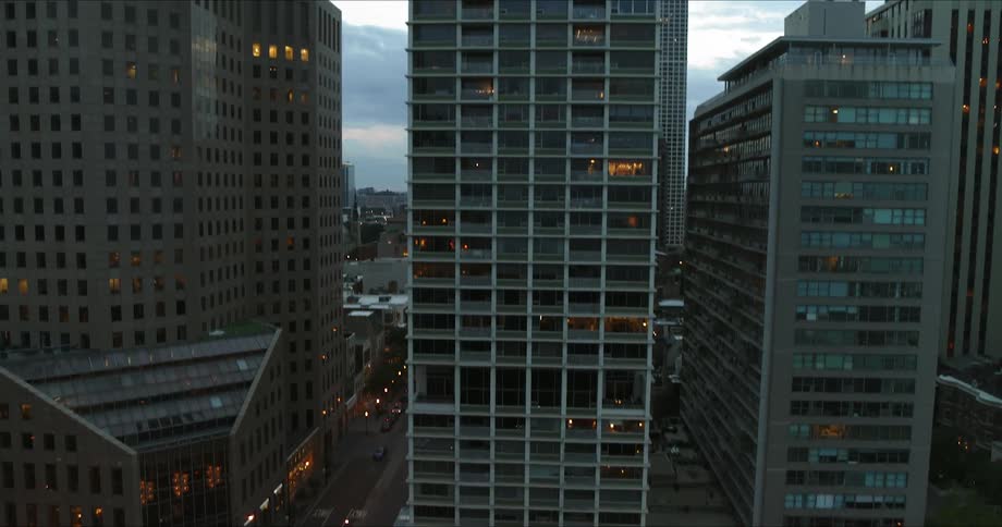 Cars travel on the street below tall city buildings with some lights glowing in windows