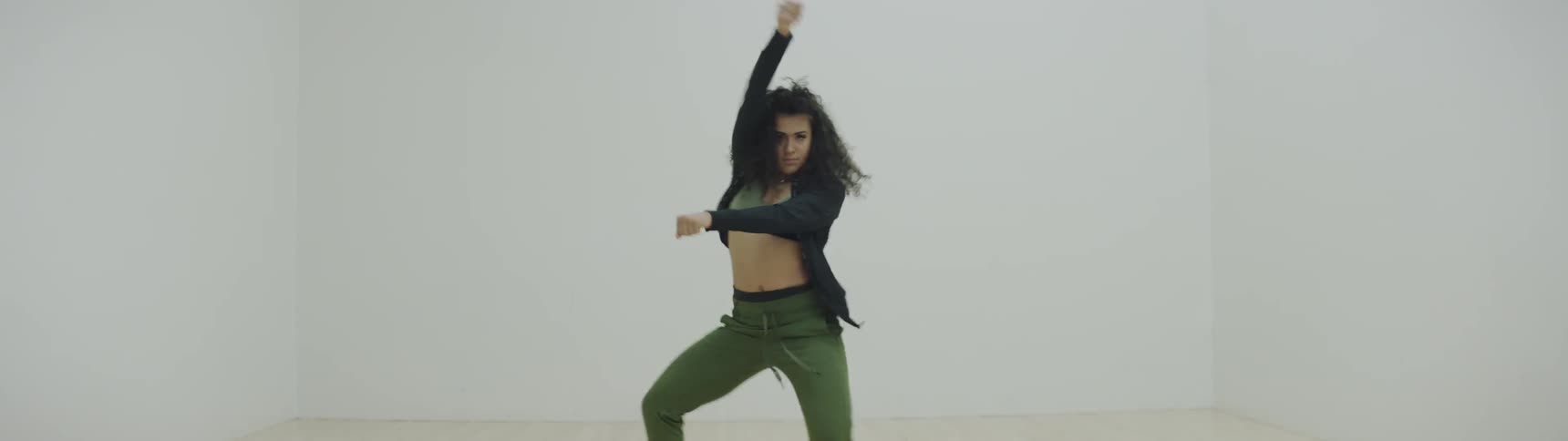 A young woman with dark curly hair performs a modern hip hop dance within the confines of racquetball court