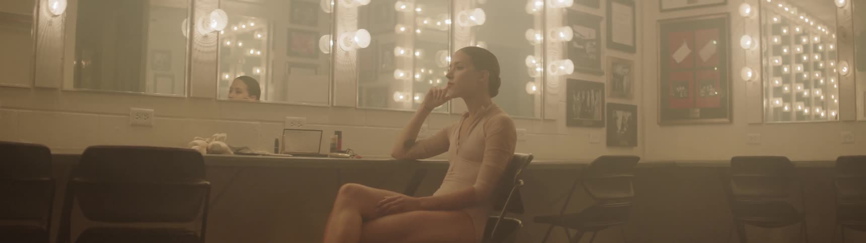 A ballerina sits in a chair at a makeup table and stares into the distance