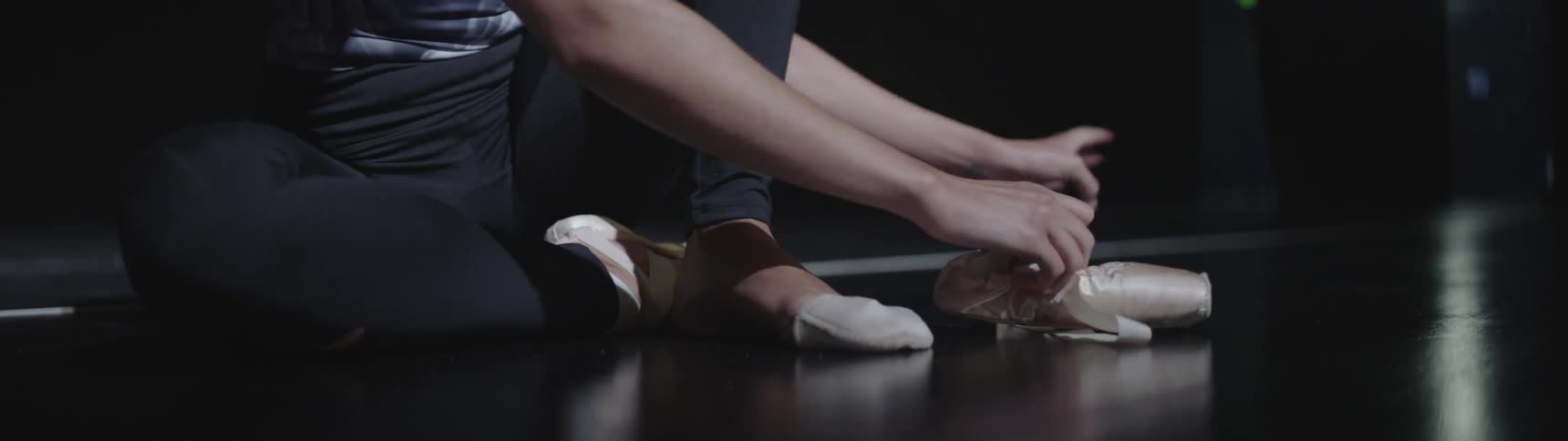 Woman wearing black leggings is sitting on the floor in a dance studio while putting on and securing her pink ballet toe shoes
