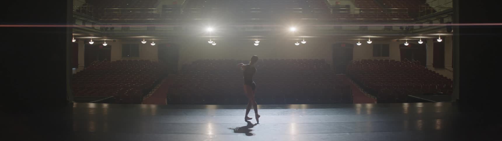 A single female ballerina dances on a large theater stage in front of an entirely empty seating area