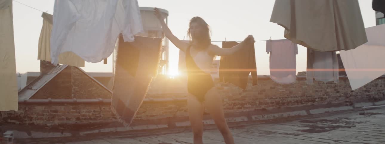 A woman dances on top of a roof with clothes hanging around while the sun sets