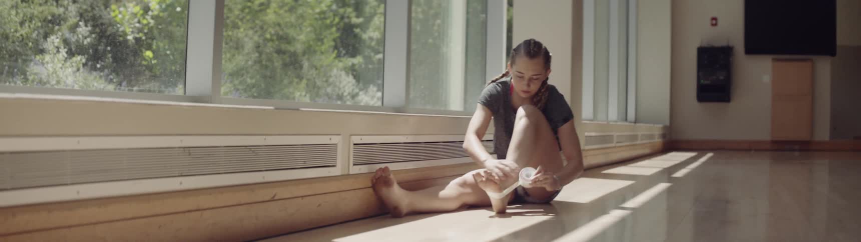 A teenage dancer sits on a studio floor and tapes her foot