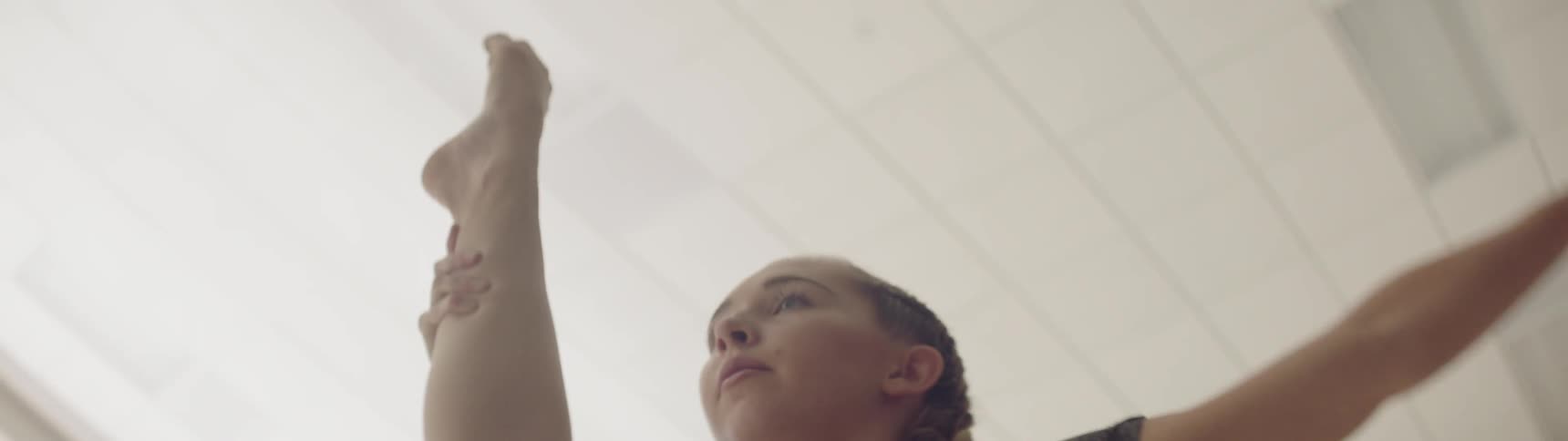 A young white woman stretches her leg above her head in a dance studio