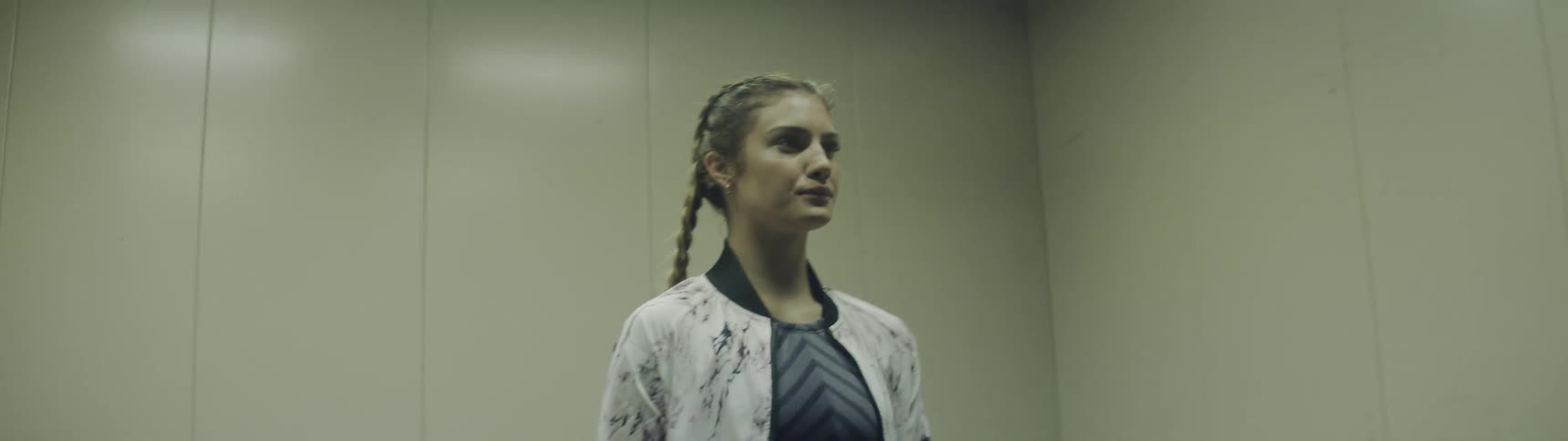 A young white woman in urban wear with braids exiting a gated lift and walking through a hallway