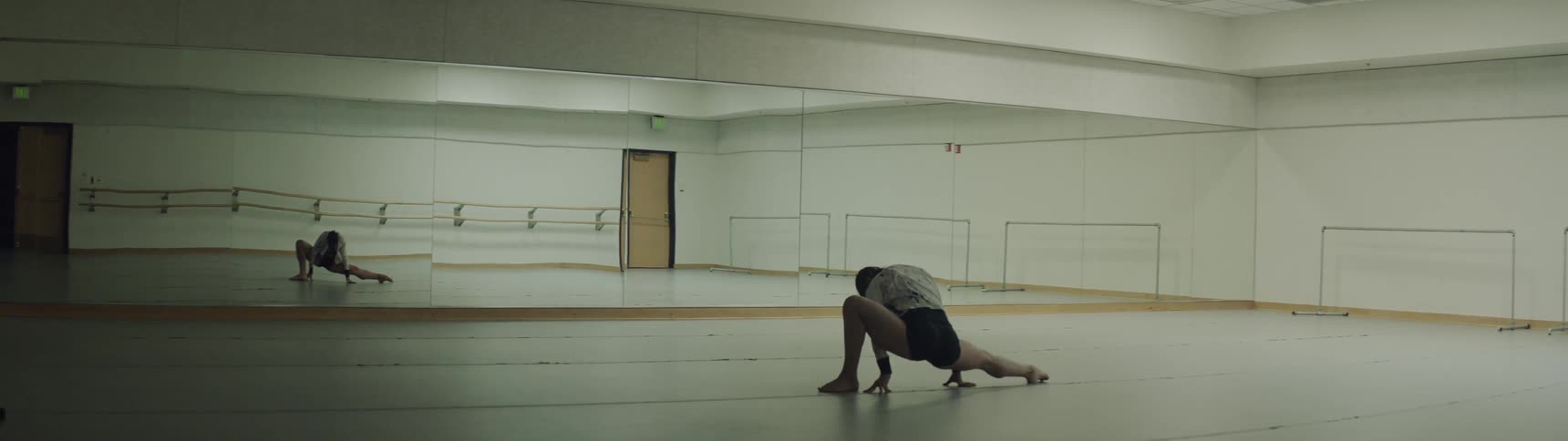 A female wearing a leotard is stretching on the floor of a dance studio in front of a wall of mirrors